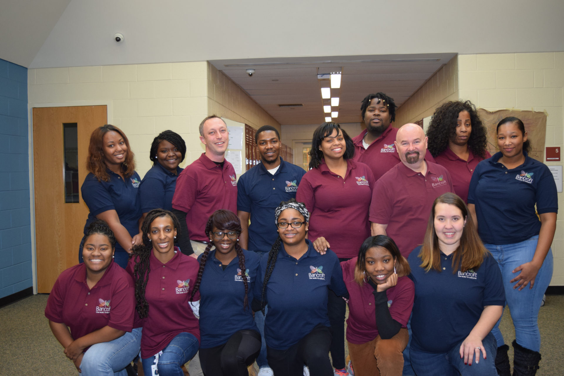 15 paraprofessionals in maroon and blue collared shirts stand and kneel in two lines smiling