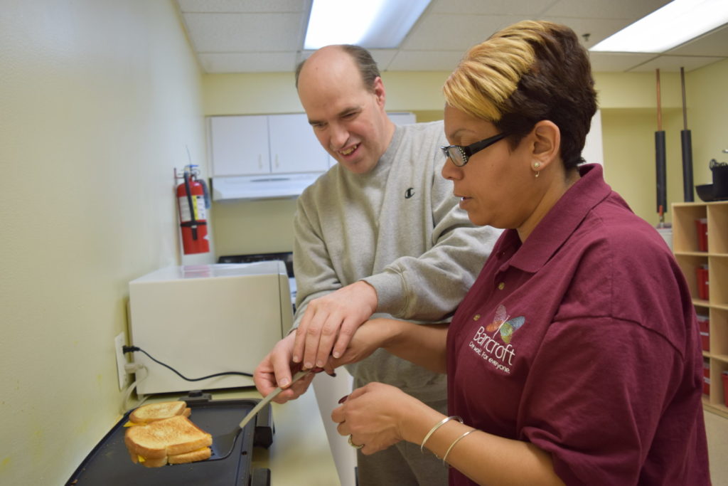 A male student in a gray crew neck and a female paraprofessional in a maroon shirt make two grilled cheese sandwiches