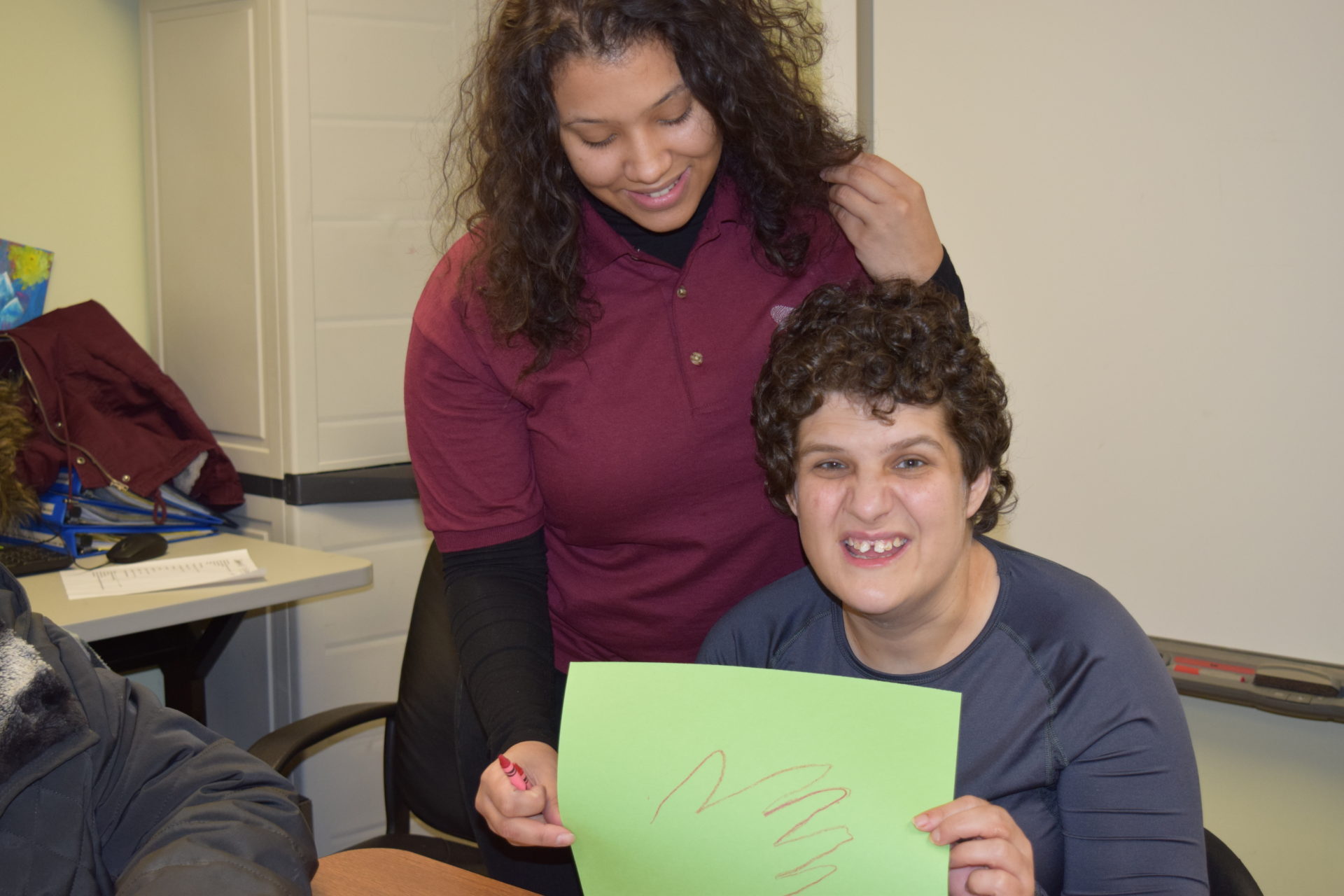 Female paraprofessional in maroon shirt with a female resident sitting at a desk in a gray shirt holding up a green paper