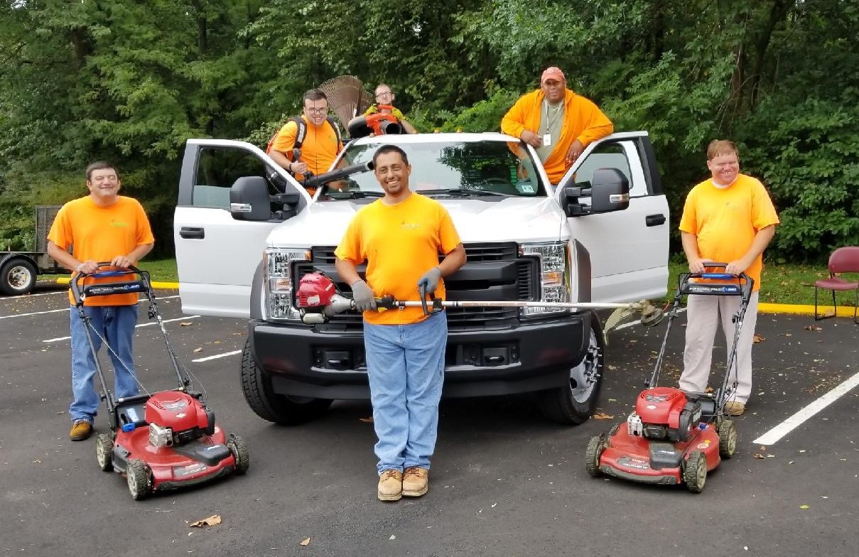 A group of vocational workers in bright orange t-shirts, standing in front of a white truck. They have lawn mowers and other landscaping equipment.