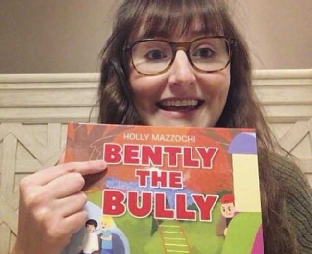 Holly Mazzochi, a light-skinned woman with brown hair and glasses holds her book "Bently the Bully" and smiles