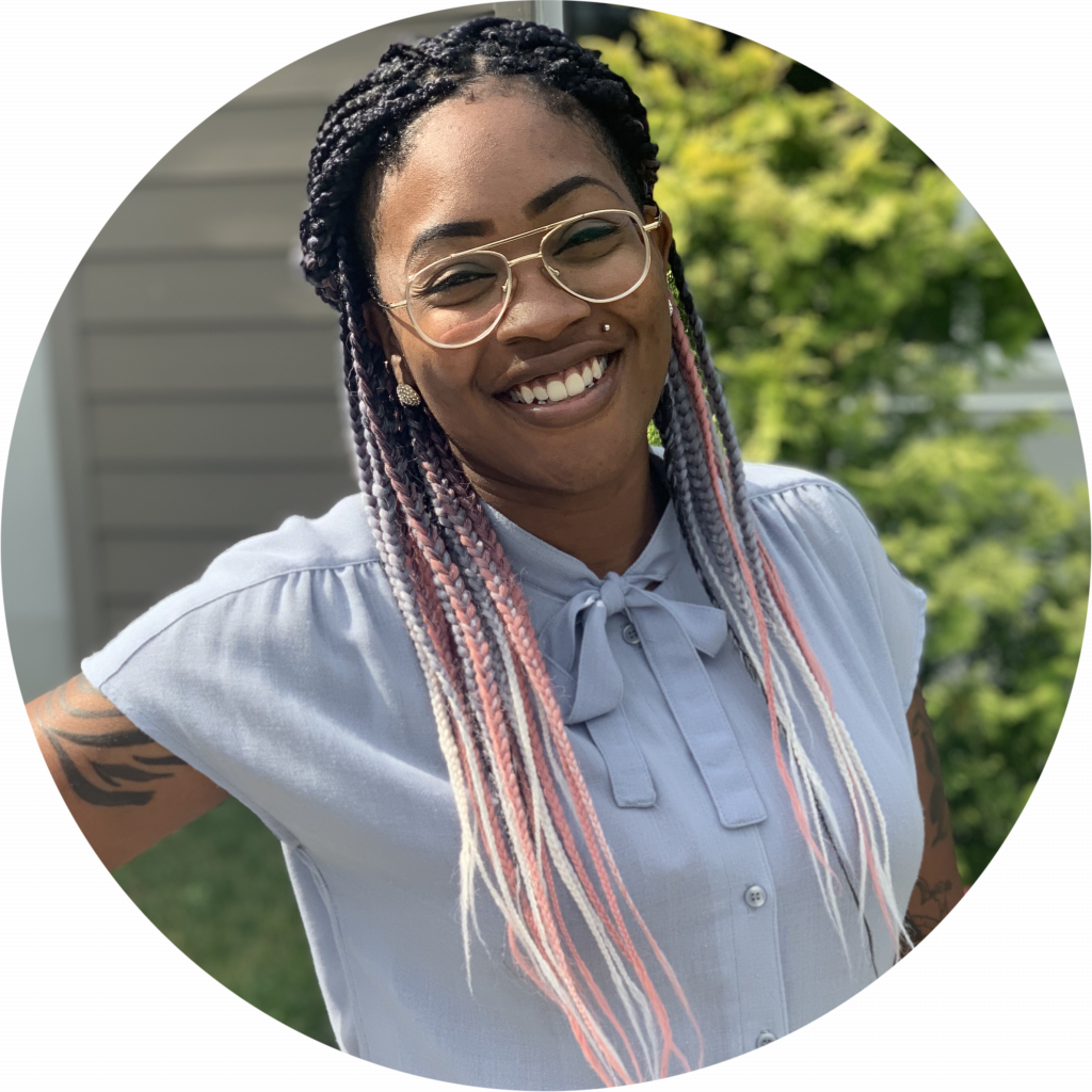 Female Direct Support Professional smiling and posing for the camera. She is wearing a blue short sleeve collared shirt and glasses. Her hair is braided and has pink at the ends.