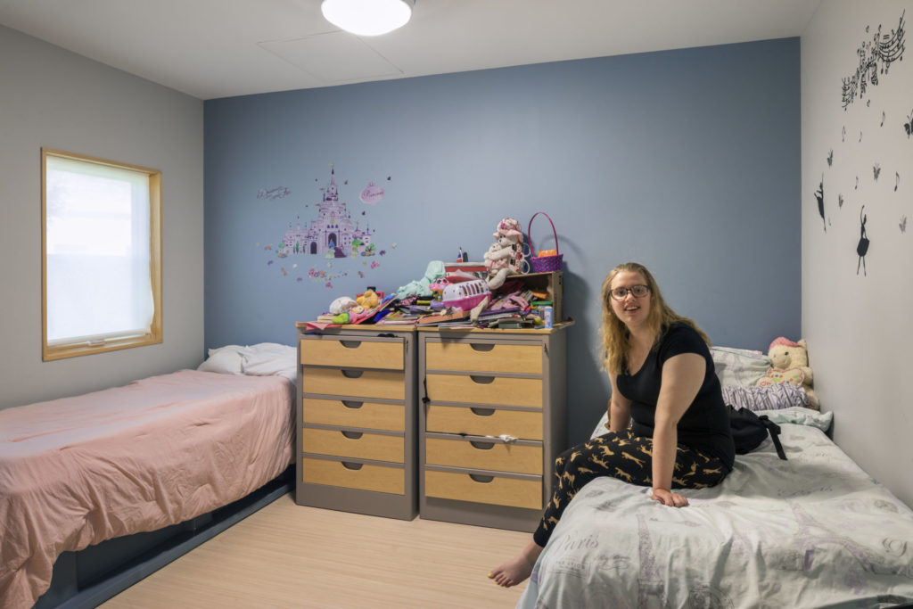 Young girl sitting in her room with a blue wall and pink bedding. Toys are stacked up behind her