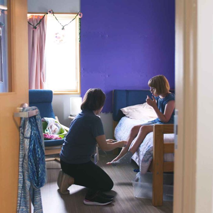 Paraprofessional helping a child put on her shoes. They are in a bedroom and the child is sitting on her bed. She has blonde short hair and a blue shirt. Her walls are purple.