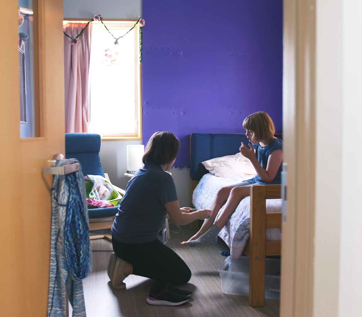Paraprofessional helping a child put on her shoes. They are in a bedroom and the child is sitting on her bed. She has blonde short hair and a blue shirt. Her walls are purple.