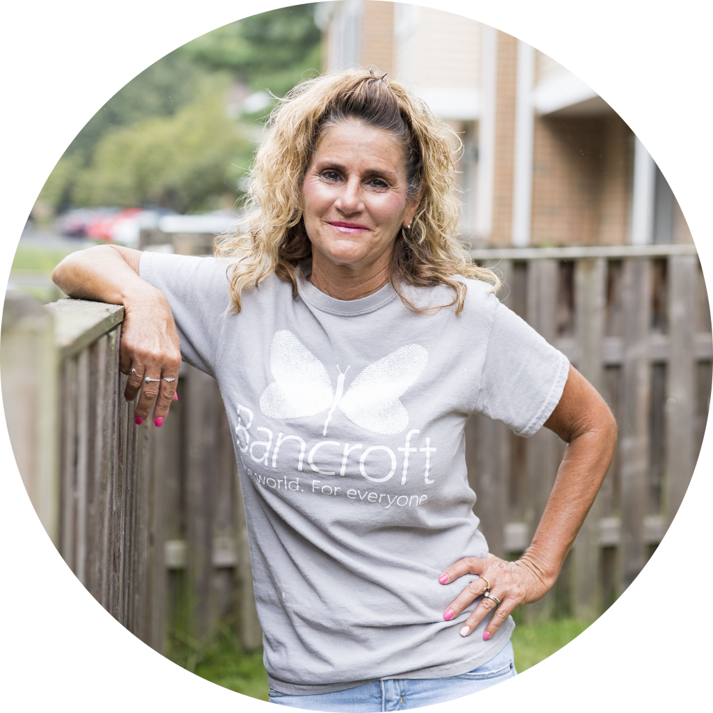 Female direct support professional standing with her arm resting on a fence post. She is wearing a grey Bancroft t-shirt. She is outside and smiling posing for the picture.