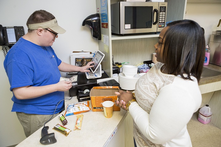 A young adult male in a blue shirt giving change to a customer. He is standing in front of a cash register. The custer is an adult female and she is wearing a while long sleeve shirt.