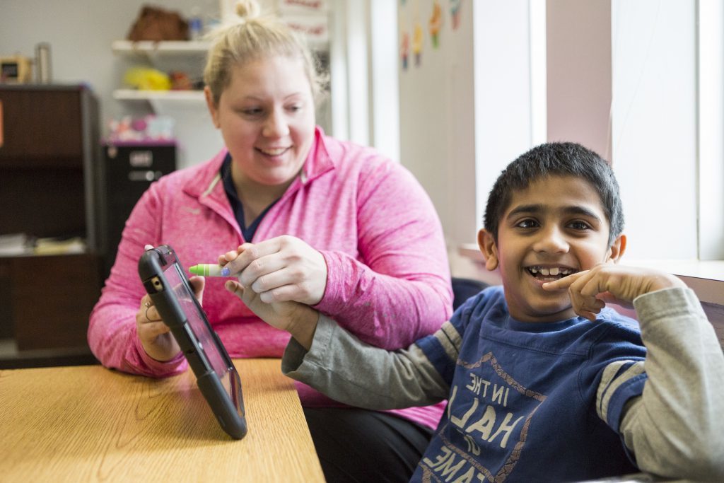 Female paraprofessional in bright pink sweatshirt sitting at a table holding an ipad with her male student. Student is smiling at the camera.