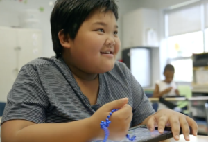 Ben; a young boy wearing a striped t-shirt sits at a desk playing with a tablet and smiling