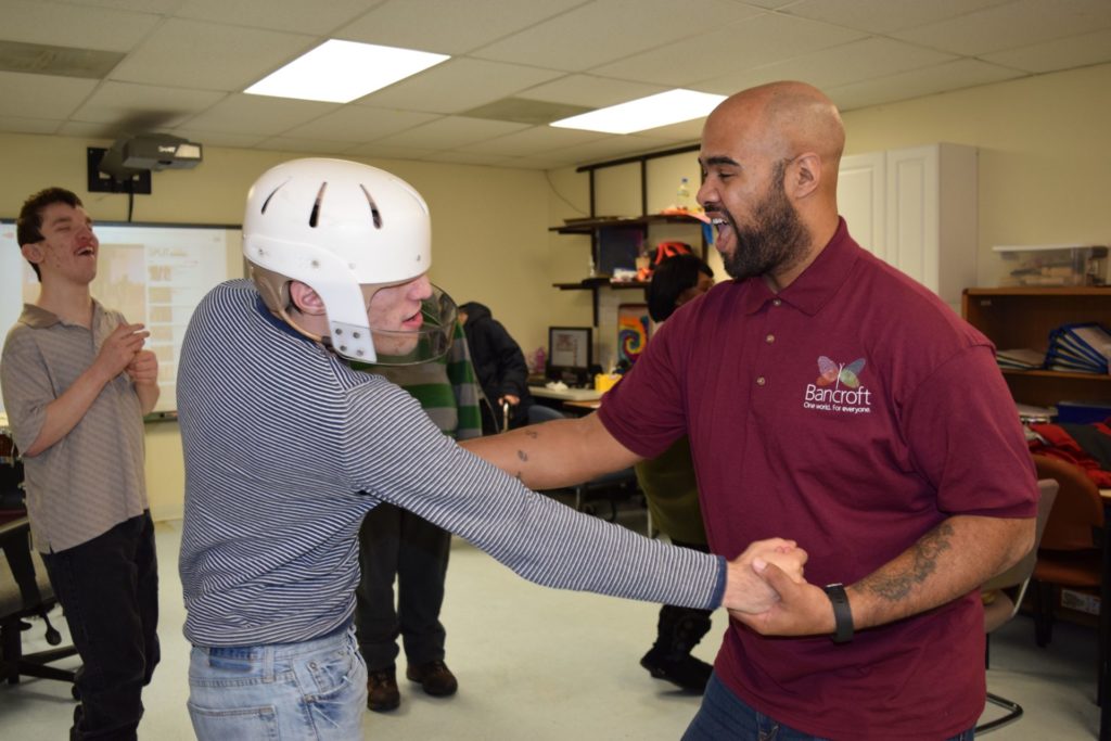 A paraprofessional in a maroon shirt and a student in a striped shirt, jeans, and helmet play together and hold hands