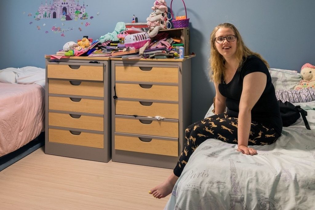 Young girl sitting in her room with a blue wall and pink bedding. Toys are stacked up behind her