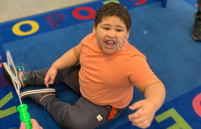 A preschool boy in an orange shirt smioling while touching bubbles that his teacher is blowing.