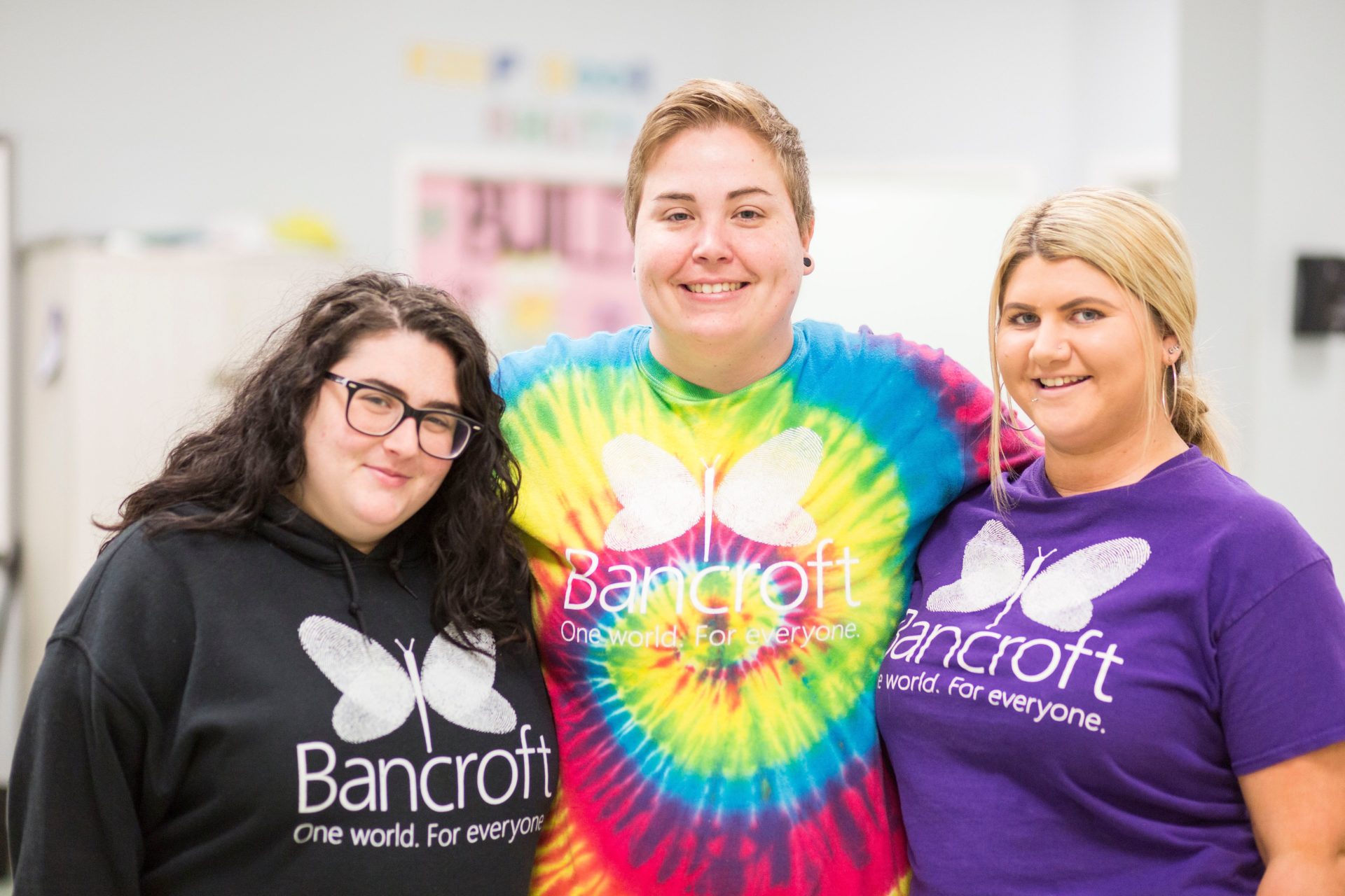 Three light-skinned females wearing bancroft t-shirts smiling next to each other