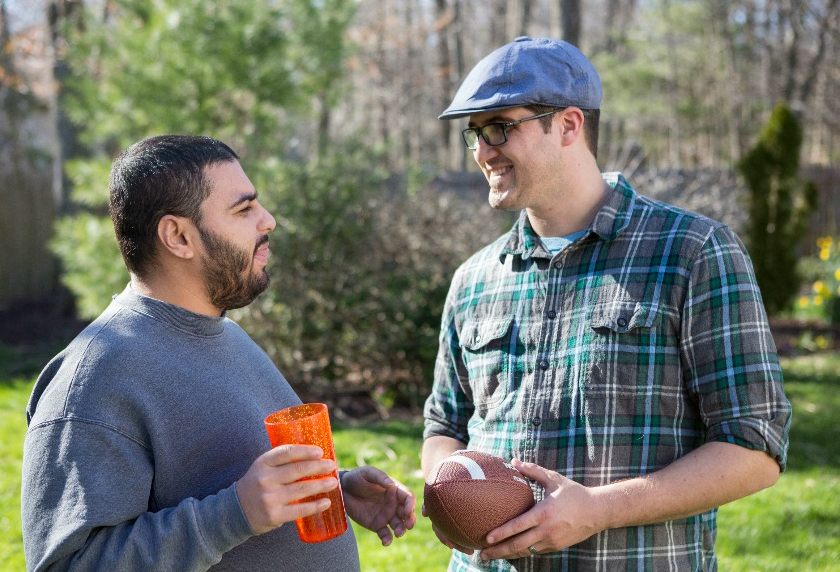 Light-skinned man in a plaid shirt and blue hat holds a football and smiles with a man in a gray sweatshirt