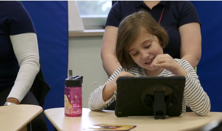 Sophia, a young girl with brown hair and a cream top, sits at a desk with paraprofessionals on a computer