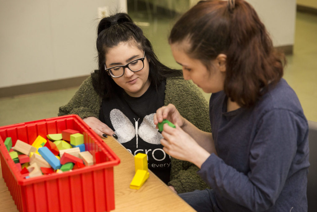 ABA Specialist working with a young girl. They are sitting at a desk and the girl has different colored blocks in her hands and next to her.