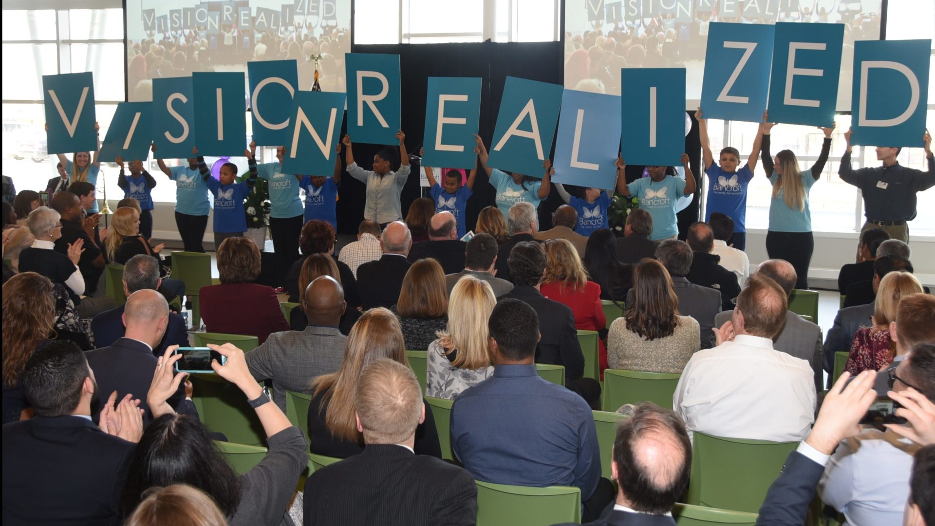 A group of young kids standing in front of a crowd holding up a sign that says "Vision Realized"