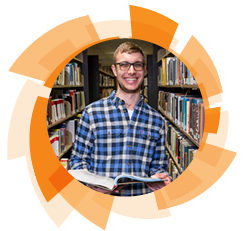 An adult male wearing a blue long sleeve button down shirt smiling at the camera. He is standing in a library holding a book.