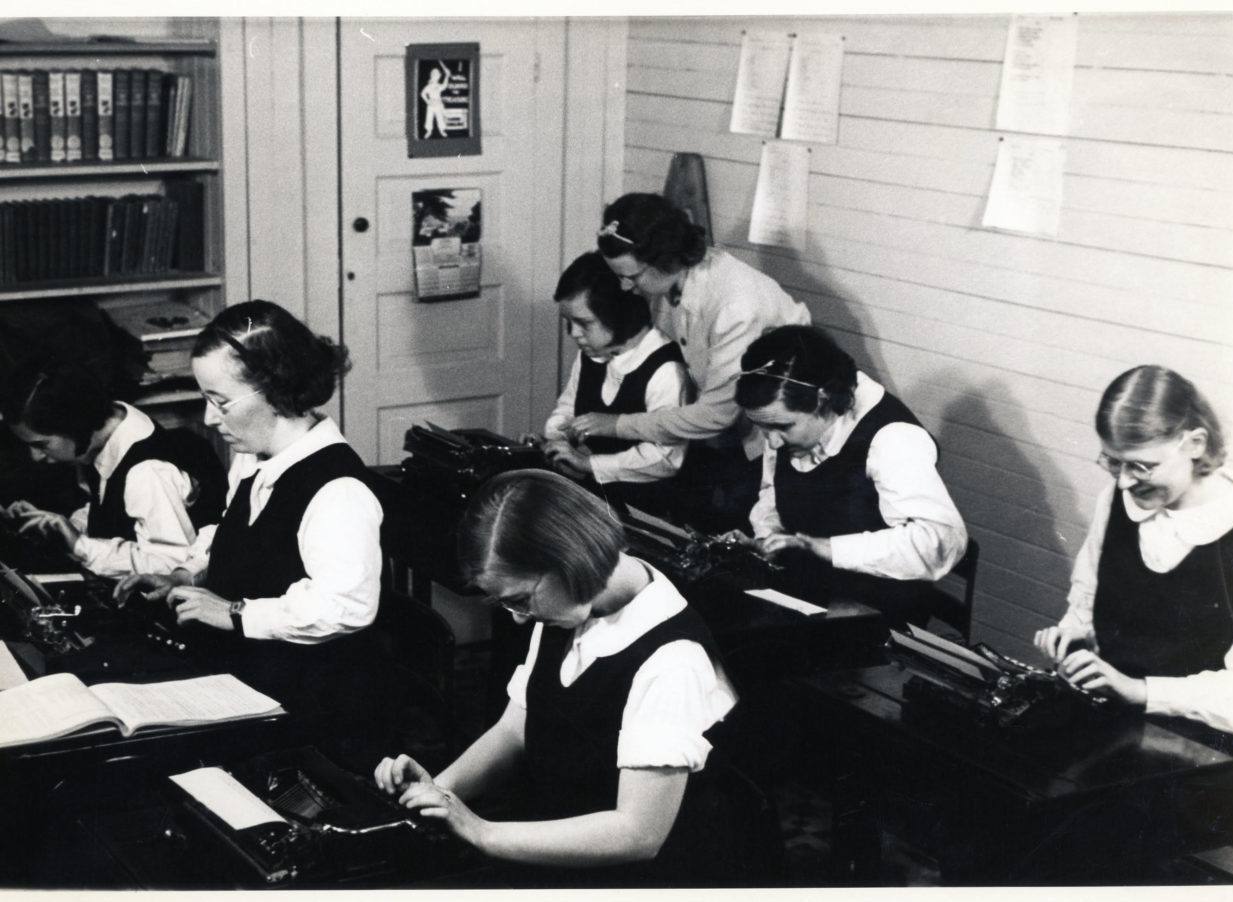 a black and white photo of six students typing on typewriters while a female teacher helps them