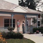 two people stand on the porch of a small yellow house with blue details sitting on a green lawn with yellow flowers