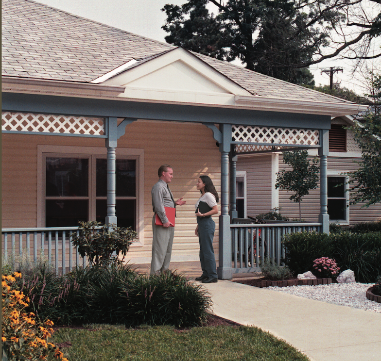 two people stand on the porch of a small yellow house with blue details sitting on a green lawn with yellow flowers