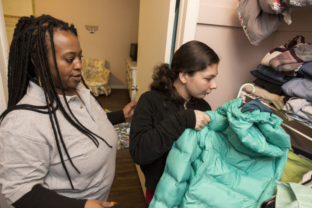 Female paraprofessional helping a young woman put her green winter jacket in the closet.
