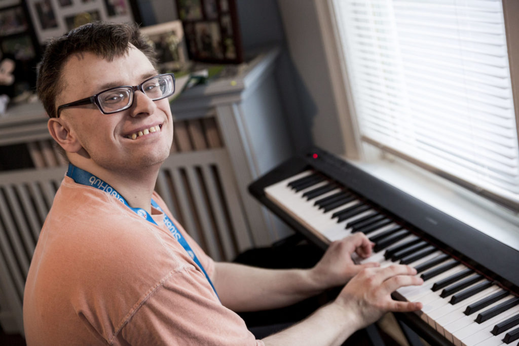An adult male playing the keyboard and smiling at the camera. He is sitting in the living room of his group home.