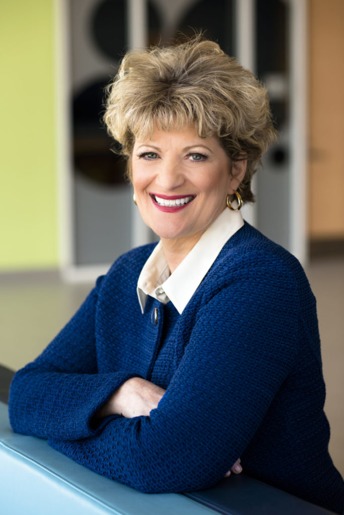 Light-skinned woman sitting with her arms crossed, smiling at the camera. She is wearing a navy blue jacket.