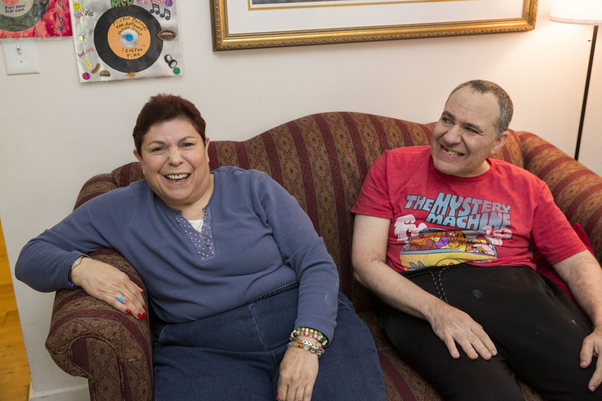 two persons served, a female in a blue shirt with short brown hair and a male in a red shirt with short hair, sit on a red couch laughing