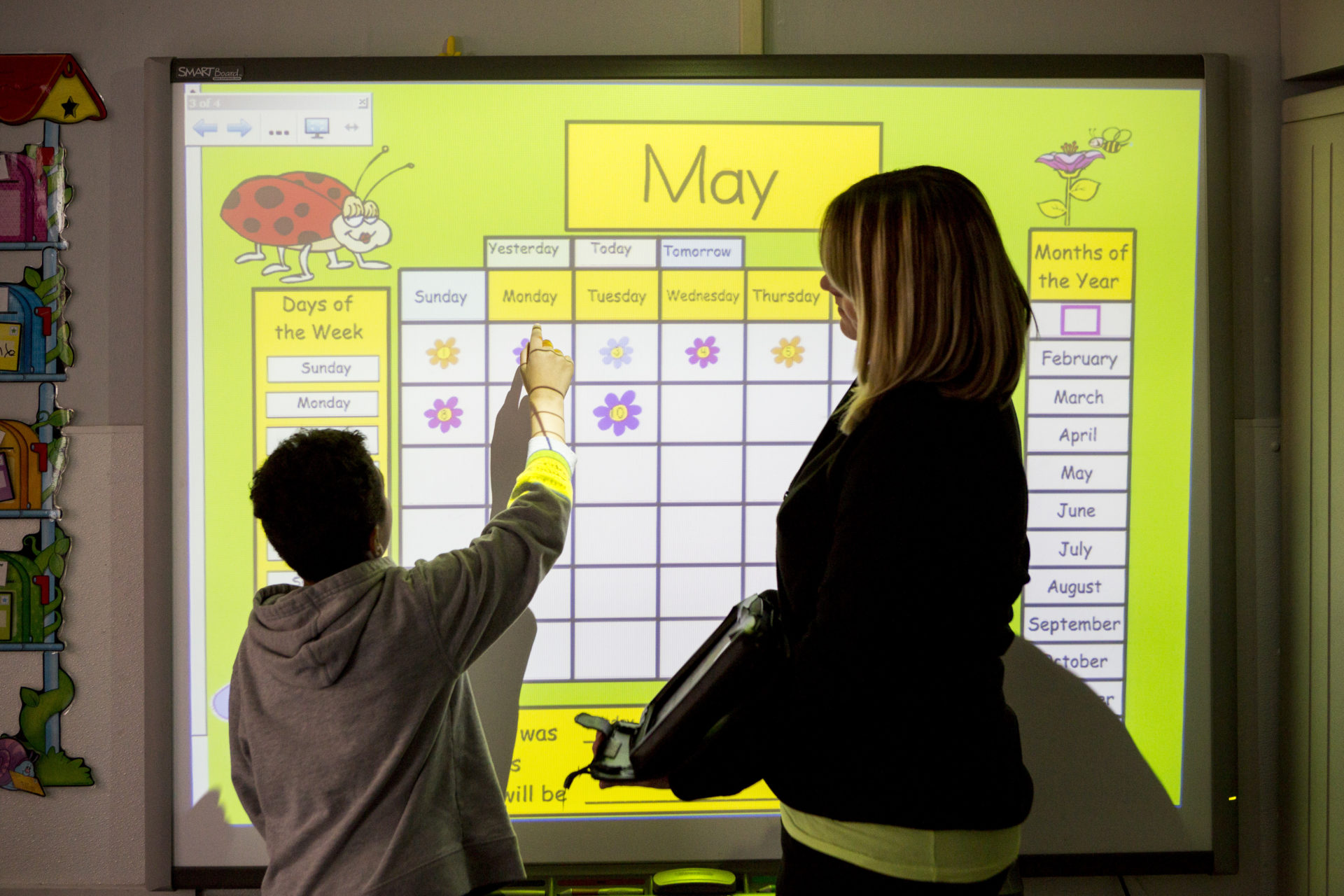 A young boy and his teacher stand in front of the smart board. They are point at a calendar for the month of May.