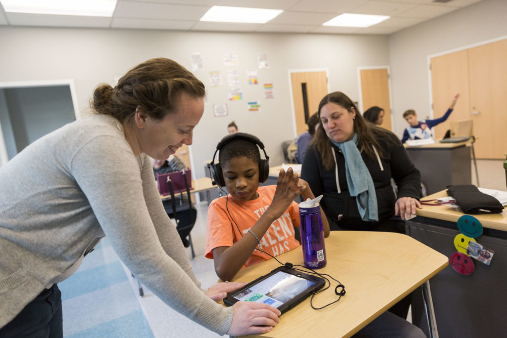 a classroom with children and paraprofessionals focusing on a boy wearing headphones using an iPad with a female paraprofessional