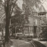 black and white photo of an old victorian home with a porch and cone roof sitting on a lawn with a large tree