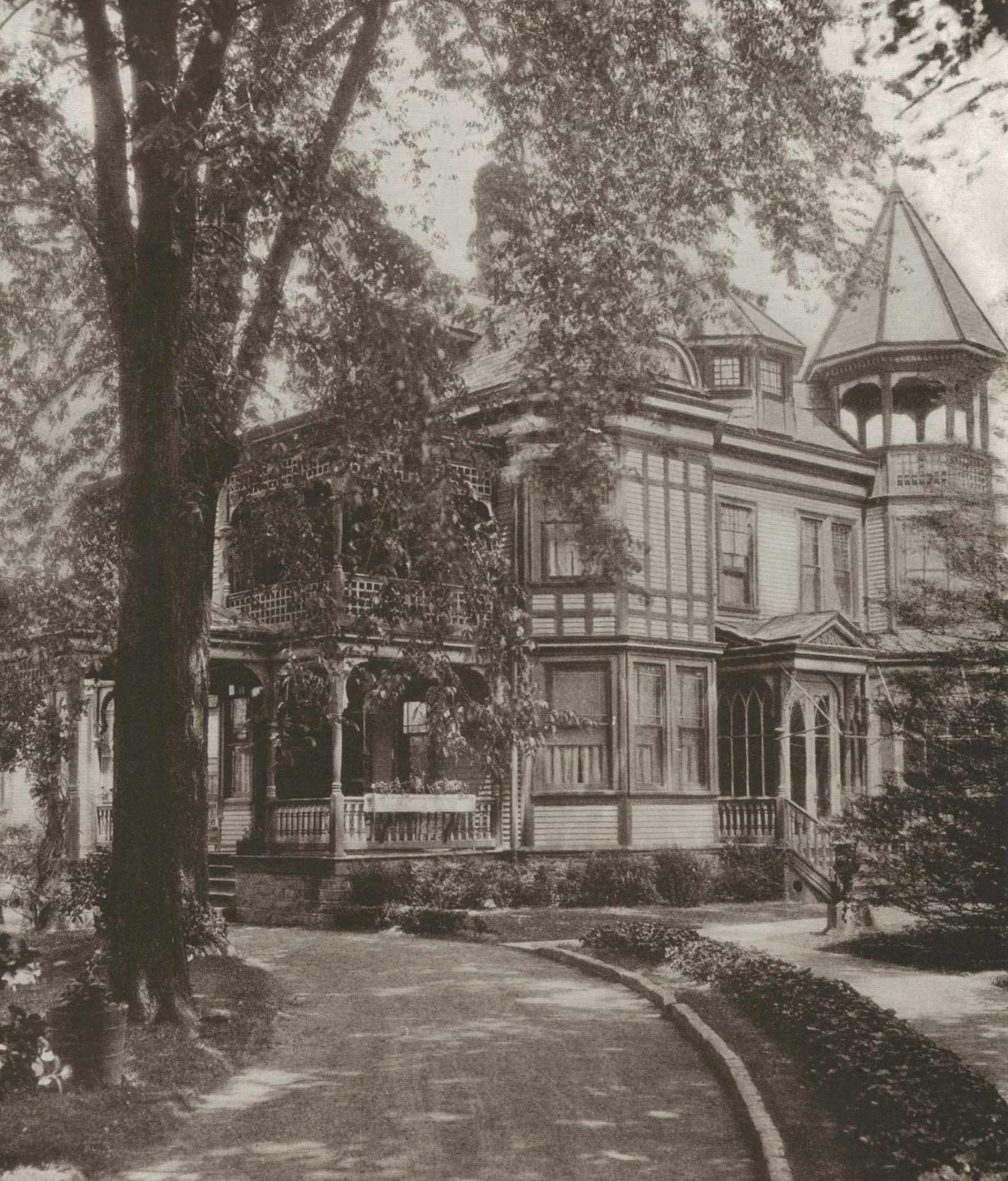 black and white photo of an old victorian home with a porch and cone roof sitting on a lawn with a large tree