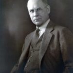 a black and white photo of a man with glasses wearing a suit and tie