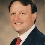 George Niemann, a light-skinned male with short black hair wearing a blue suit with a red tie, smiles