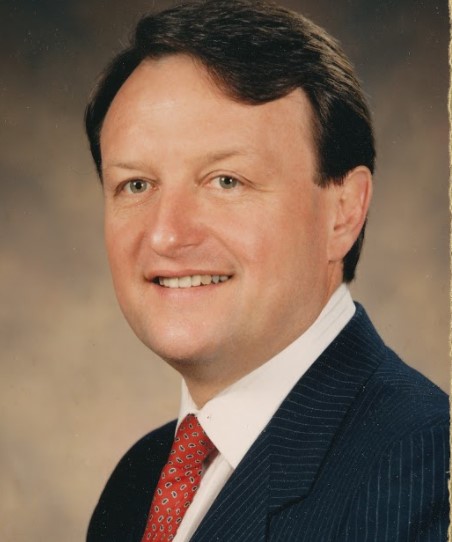 George Niemann, a light-skinned male with short black hair wearing a blue suit with a red tie, smiles