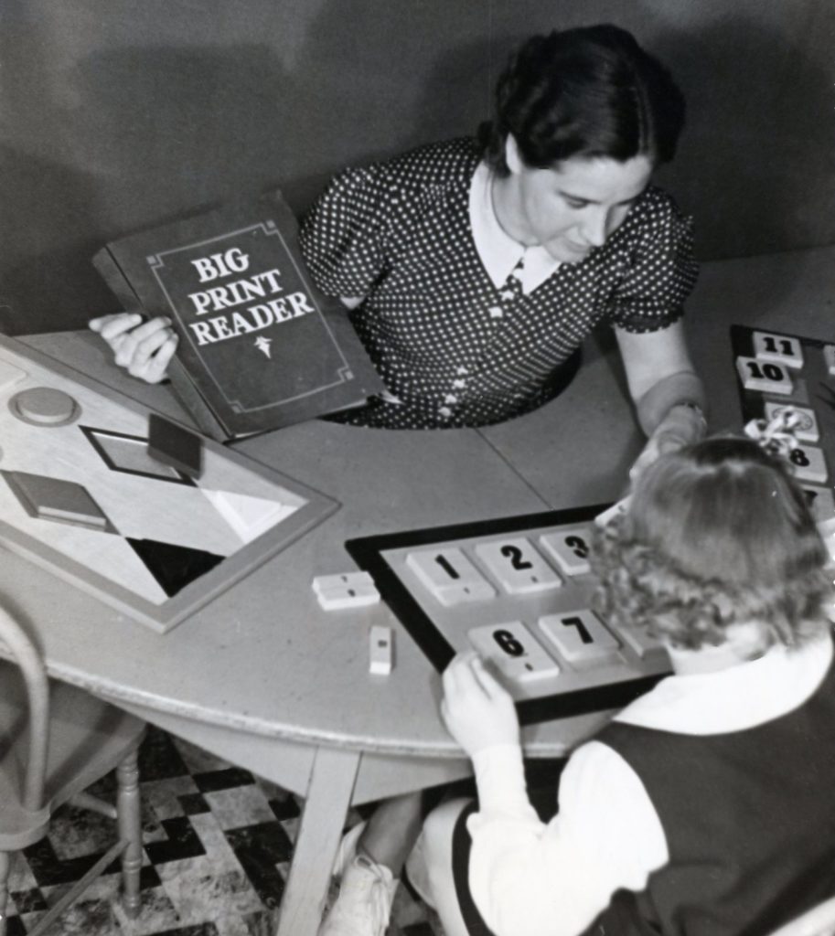 A black and white photo of a Bancroft teacher and student in the early 1900's. A small girl is reading a book, the female teacher is sitting across from her, pointing to a workboard with numbers.