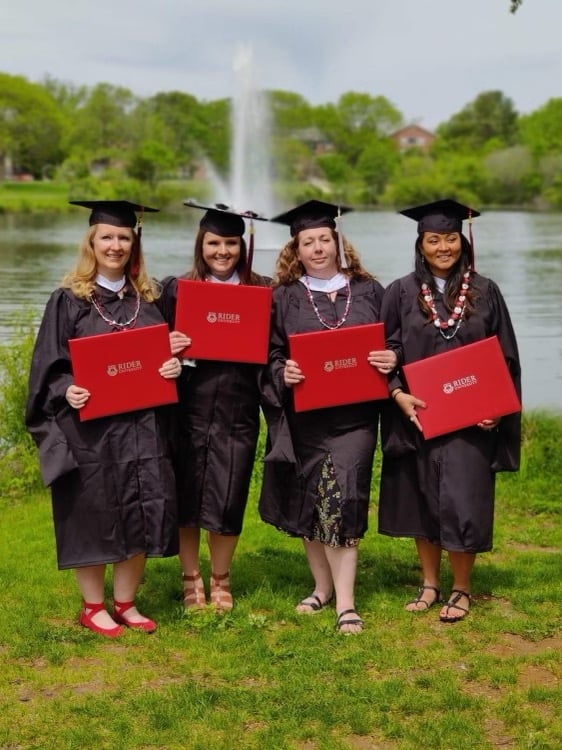 Four graduated are standing and smiling for the camera. They are in black graduation gowns and are holding red diplomas. They are standing in front of a fountain and lake.