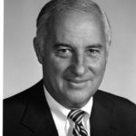 a black and white photo of a light-skinned man wearing a suit and tie, smiling