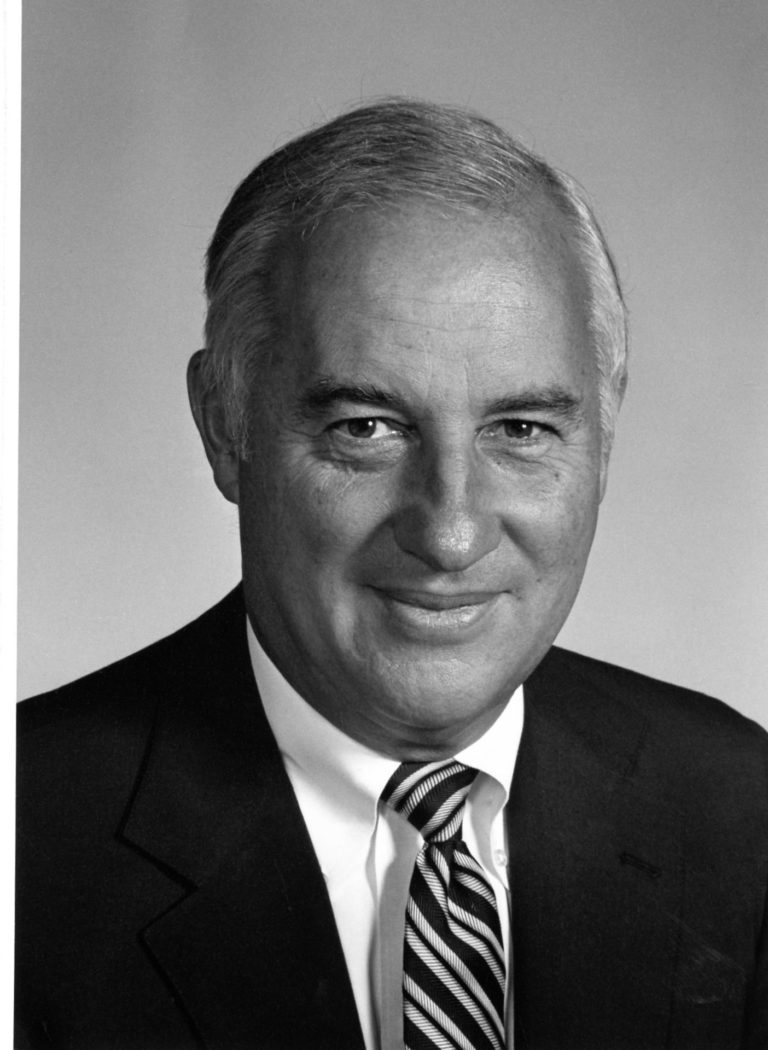 a black and white photo of a light-skinned man wearing a suit and tie, smiling