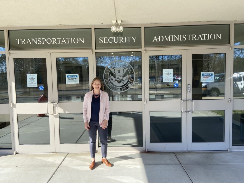 a light-skinned woman with blonde hair wearing a pink suit jacket and blue pants stands in front a building with the sign "Transportation Security Administration"
