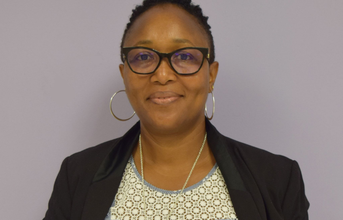 A dark skinned woman smiling at the camera. She is standing in front of a purple wall.