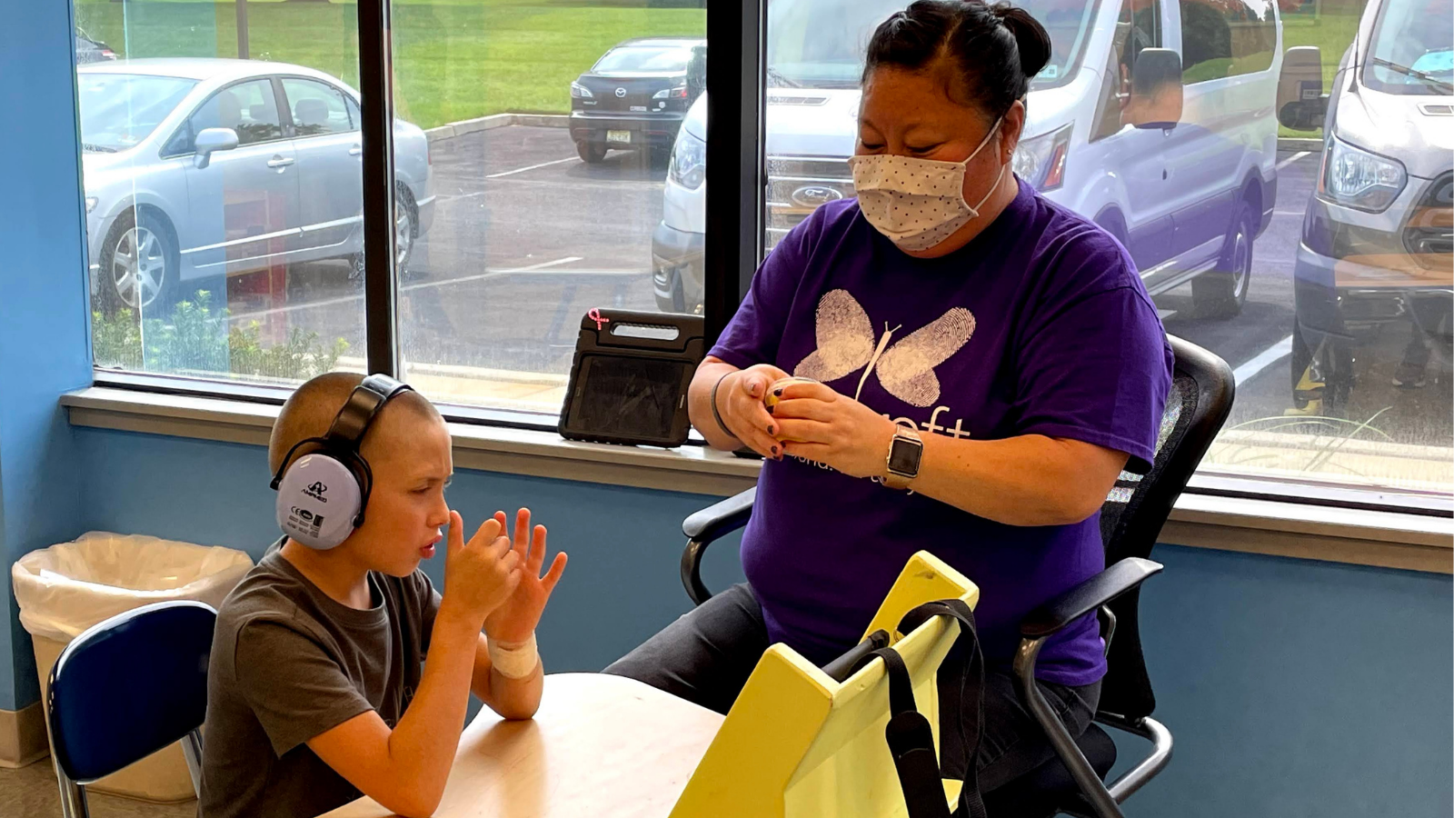 A light skinned middle aged woman sits with a preschool aged child at a table. in the classroom. They are looking at an ipad and playing with modeling dough.