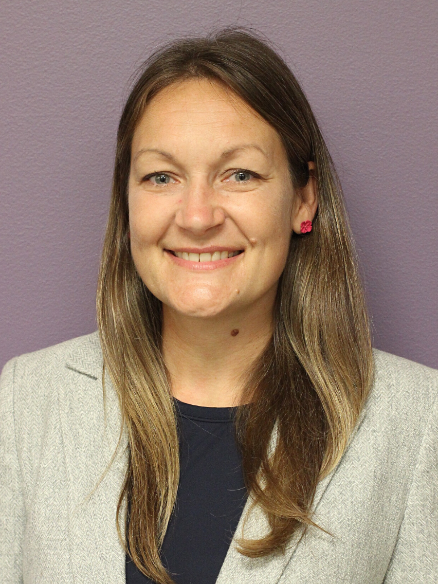 A light skinned woman stands in front of a purple wall. She has long brown hair and is smiling at the camera.