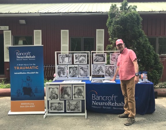 A Bancroft NeuroRehab client stands in front of a table featuring his artwork.