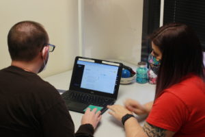 Matt Collura sits in front of his computer, Bancroft NeuroRehab Occupational Therapy Assistant sits next to him.