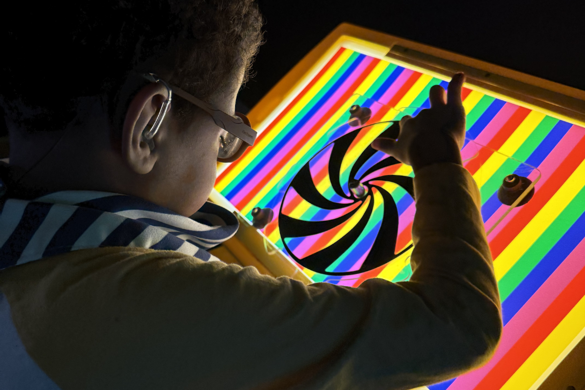 young boy sitting near a rainbow light board in a sensory room