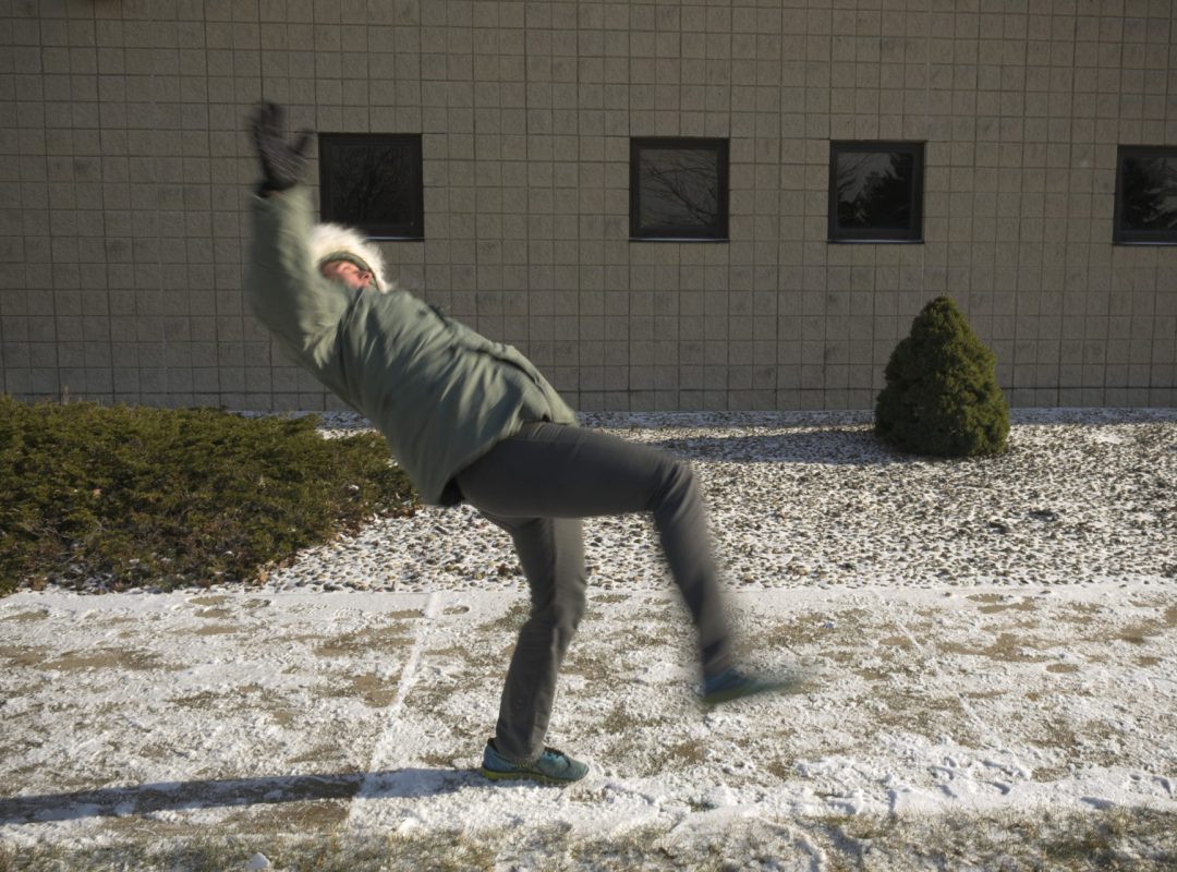 A person in a green jacket and gray pants slips on an icy sidewalk