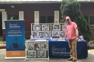 A Bancroft NeuroRehab client stands in front of a table featuring his artwork.