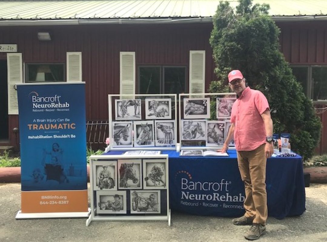 A Bancroft NeuroRehab client stands in front of a table featuring his artwork.